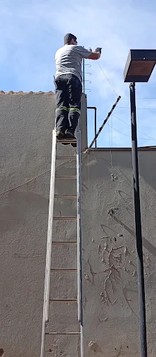Técnico em escada instalando hastes de cerca elétrica em um muro alto