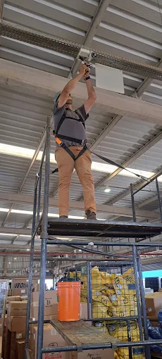 Técnico com cinto de segurança instalando uma câmera na estrutura do telhado de um galpão